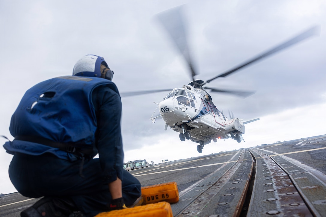A sailor in a blue vest and helmet kneels on the flight deck of a ship as a helicopter lifts off in the background on a cloudy day.