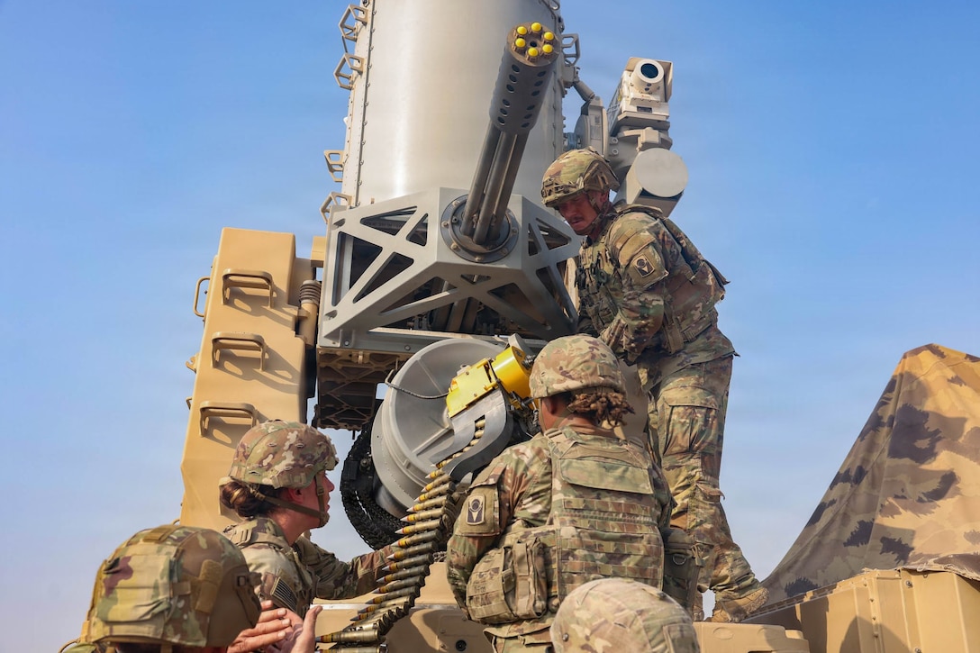 Five soldiers wearing camouflage uniforms and helmets load a belt of ammunition into a Gatling gun mounted on a platform during daytime.