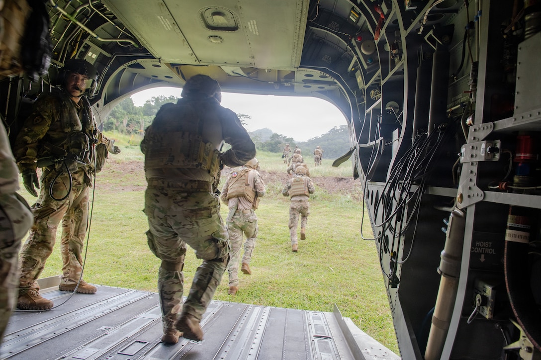 A group of about 10 service members wearing camouflage uniforms, helmets, and tactical vests exit from the tailgate of a helicopter into a field during daytime.
