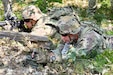 A man wearing a camouflage military uniform lies on the ground in the forest while placing a simulated explosive as another man in similar attire aims a rifle.