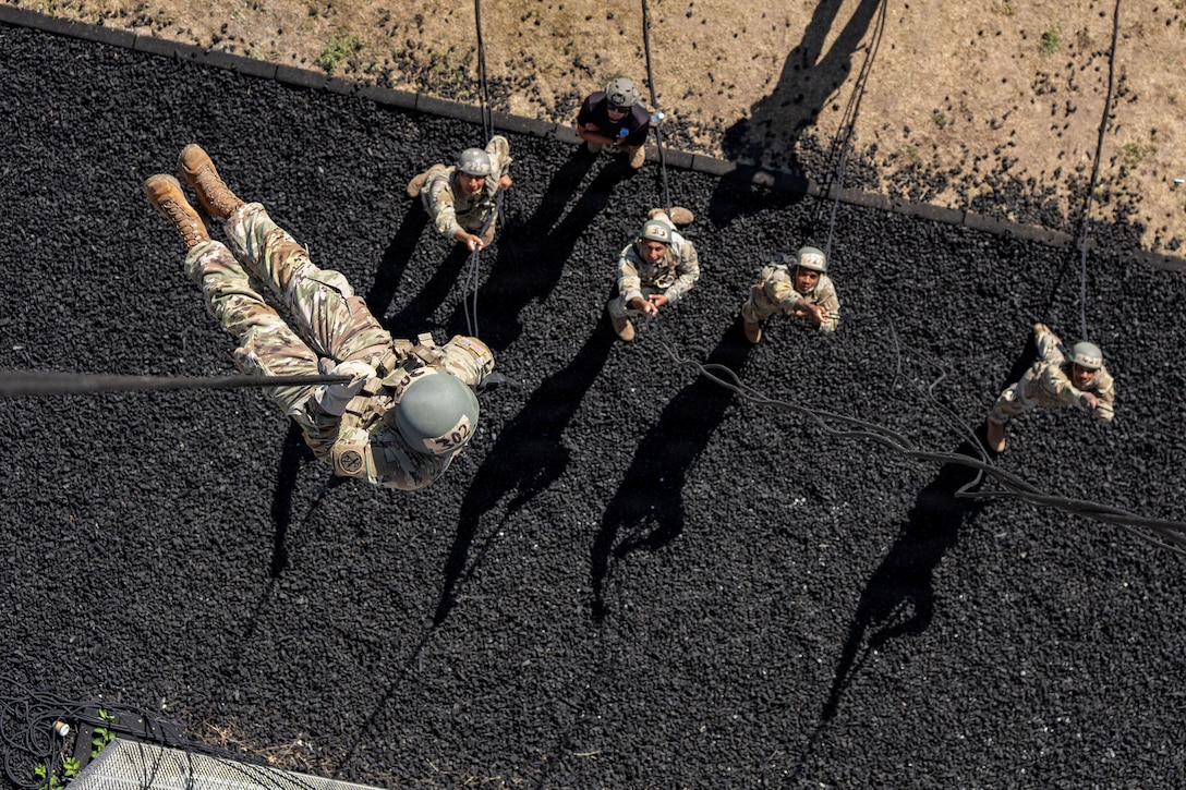 A soldier in a camouflage uniform and helmet hangs from a rope as four others standing on the ground hold ropes and another one watches.