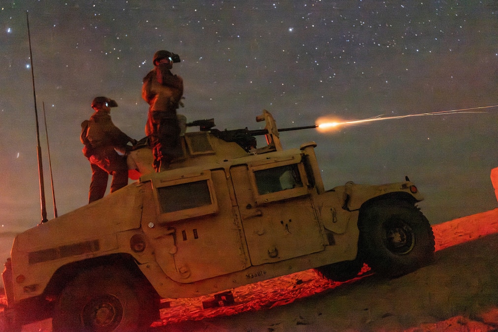 Two Marines wearing night-vision equipment stand on a military vehicle as another fires a mounted machine gun, making a flash against dark sky.
