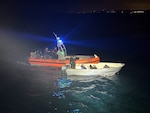 A Coast Guard small boat is seen with its blue light flashing; floating alongside a smaller panga-style vessel. Two Coast Guardsman are aboard the panga while the passengers of the vessel sit towards the bow. One of the passengers has his hands up.