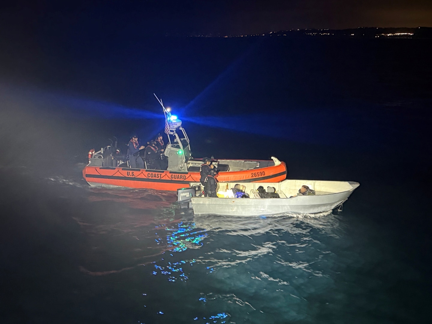A Coast Guard small boat is seen with its blue light flashing; floating alongside a smaller panga-style vessel. Two Coast Guardsman are aboard the panga while the passengers of the vessel sit towards the bow. One of the passengers has his hands up.