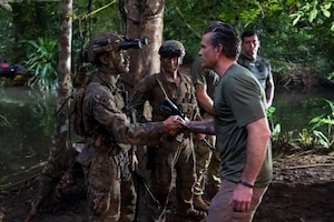 In a jungle environment, a man in casual attire shakes hands with a man in a camouflage military uniform and night vision equipment on his helmet while people in similar attire observe.