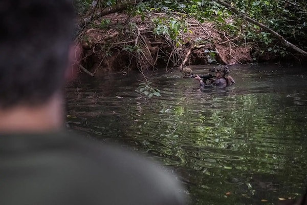 A rear-facing man observes two people in camouflage military uniforms as they move through water; one man carries a rifle.