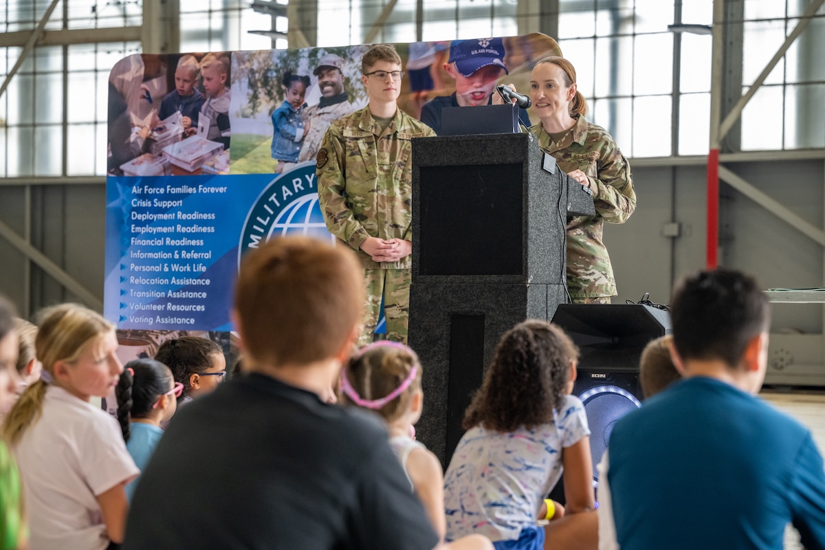 A woman, wearing camouflage military uniform, stands behind a podium and microphone in front of a group of children
