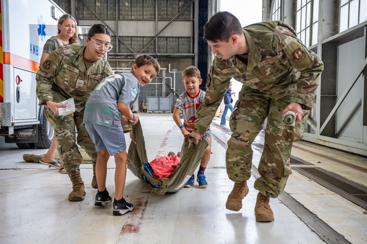 A woman and man, wearing camouflage military uniforms, help two young boys drag a tarp with carrying another child