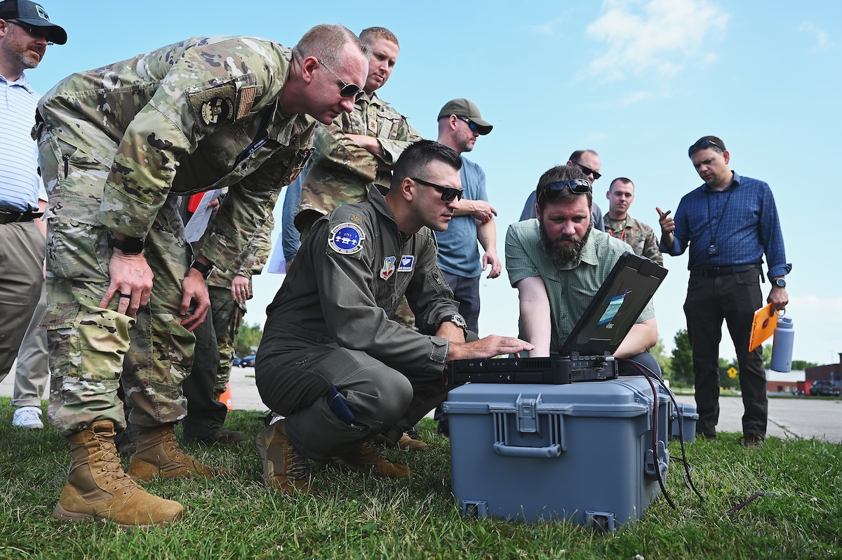 Men in Air Force uniforms and business attire stand in field on sunny day, looking at a black laptop
