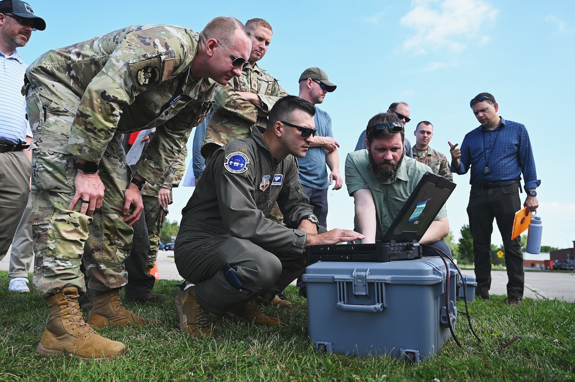 Men in Air Force uniforms and business attire stand in field on sunny day, looking at a black laptop