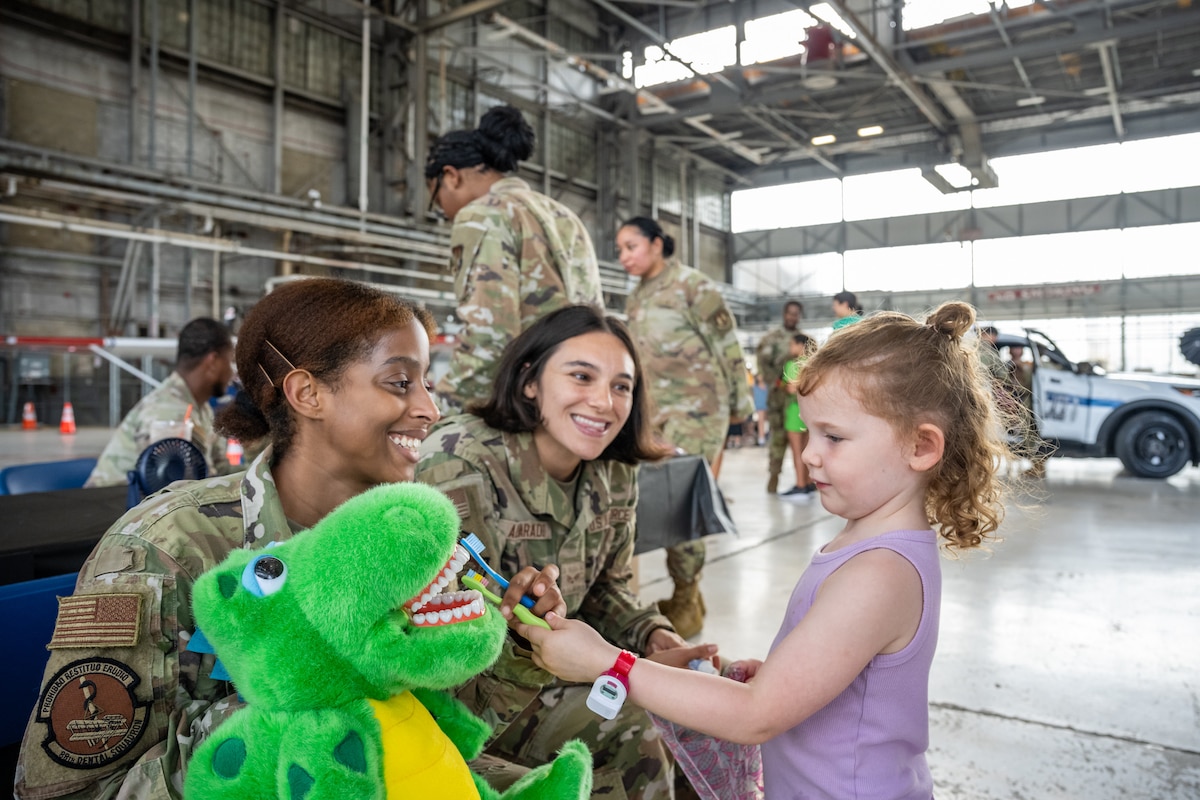 Two women, wearing camouflage military uniforms, smile as a young girl brushes the teeth of a green dinosaur plushie.