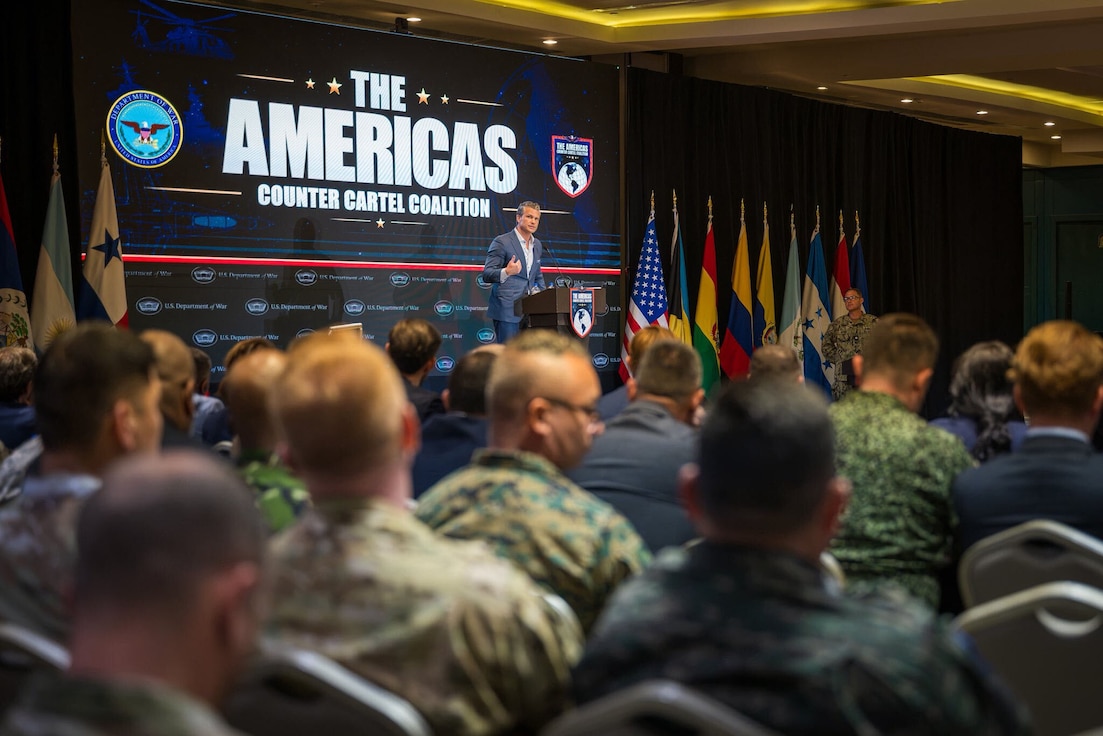 Secretary of War Pete Hegseth speaks at a lectern on a stage to a seated audience.