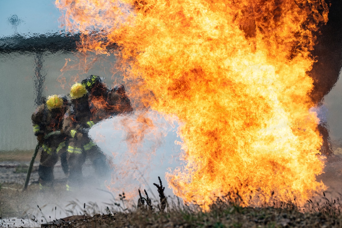 U.S. Air Force Airman 1st Class Andrew Gonzalez, Airman 1st Class Elias Villaseñor and Senior Airman Jacob Thompson, 100th Civil Engineer Squadron firefighters, extinguish a fire during the 100th CES Fire Department Rookie Academy at RAF Mildenhall, United Kingdom, July 22, 2026. The academy condenses four months of training into three weeks for new firefighters, providing consistent, high-quality instruction. (U.S. Air Force photo by Senior Airman Aidan Martínez Rosiere)