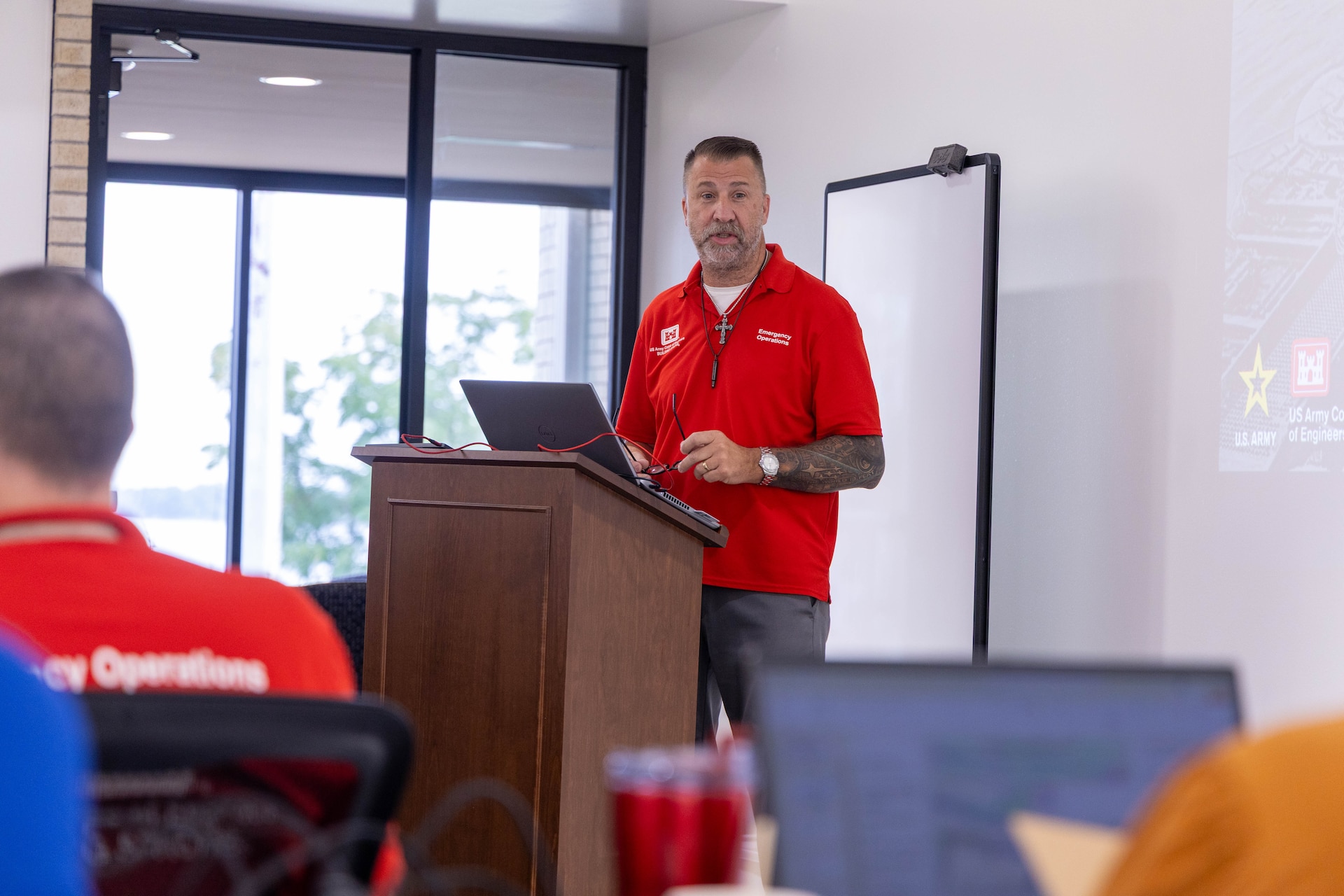 A man in a red shirt stands behind a brown podium and speaks to a room of people with a white wall in the background.