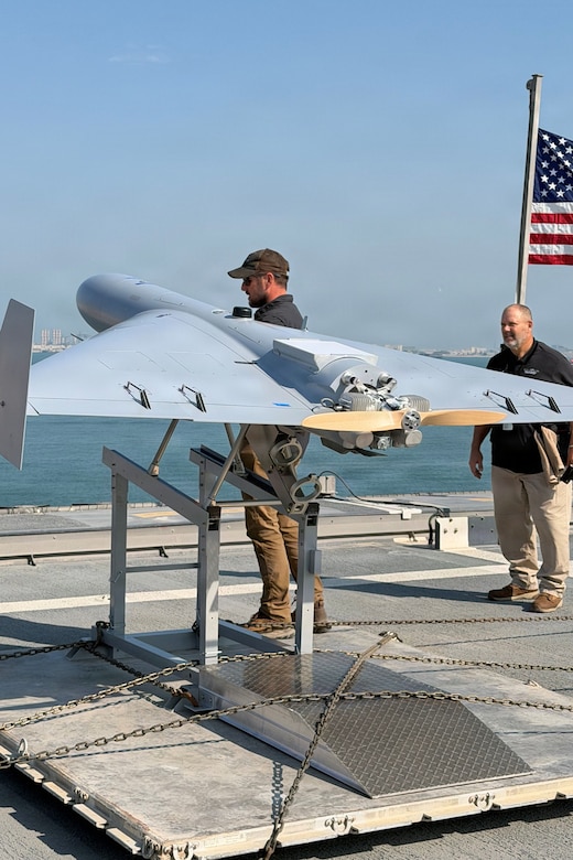 A man wearing casual attire, a baseball cap and sunglasses works on a drone as another man in similar attire looks on. They are both standing on the deck of a ship at sea.