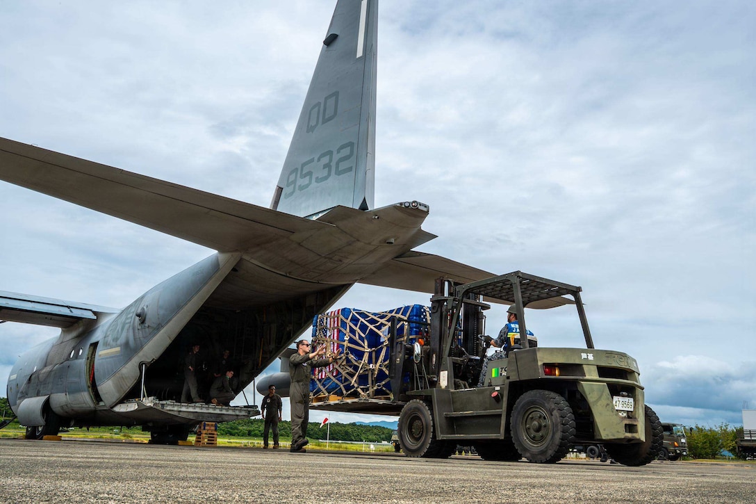 A person in a camouflage military uniform drives a forklift with a pallet of supplies on it up to a military plane parked on a tarmac as another person in a military uniform guides the forklift.