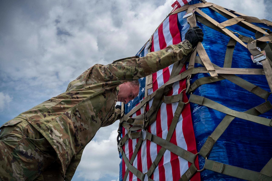 A man in a camouflage military uniform moves a pallet of supplies with an American flag attached to it.