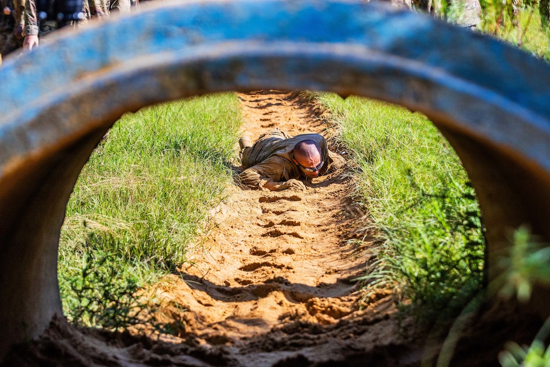 An Army trainee crawls through a muddy shallow ditch toward a tunnel structure during daytime, with partially visible people standing in the grass nearby.