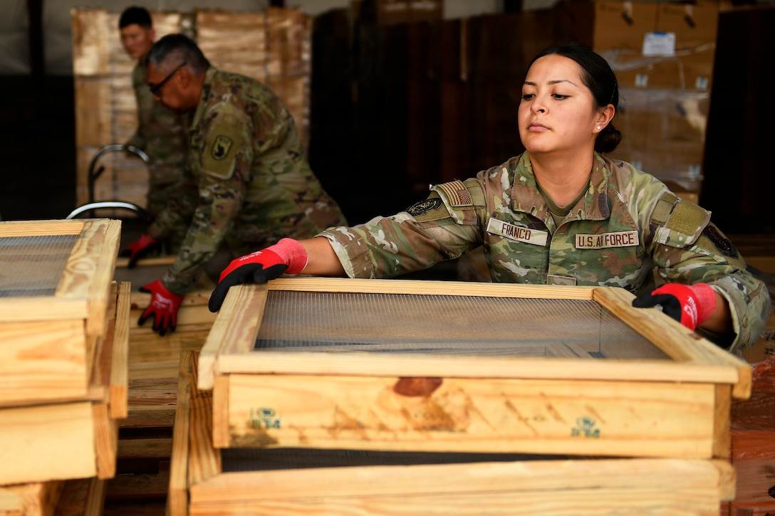 Three airmen in camouflage uniforms assemble wooden boxes in a warehouse-like setting, with boxes stacked in the background.