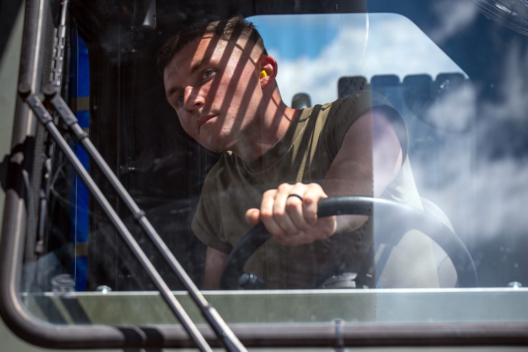An airman steers the wheel of a military vehicle during daytime, as seen through the front windshield.