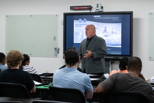 A man stands at the front of a conference room in front of a monitor speaking to a group of people seated at tables.