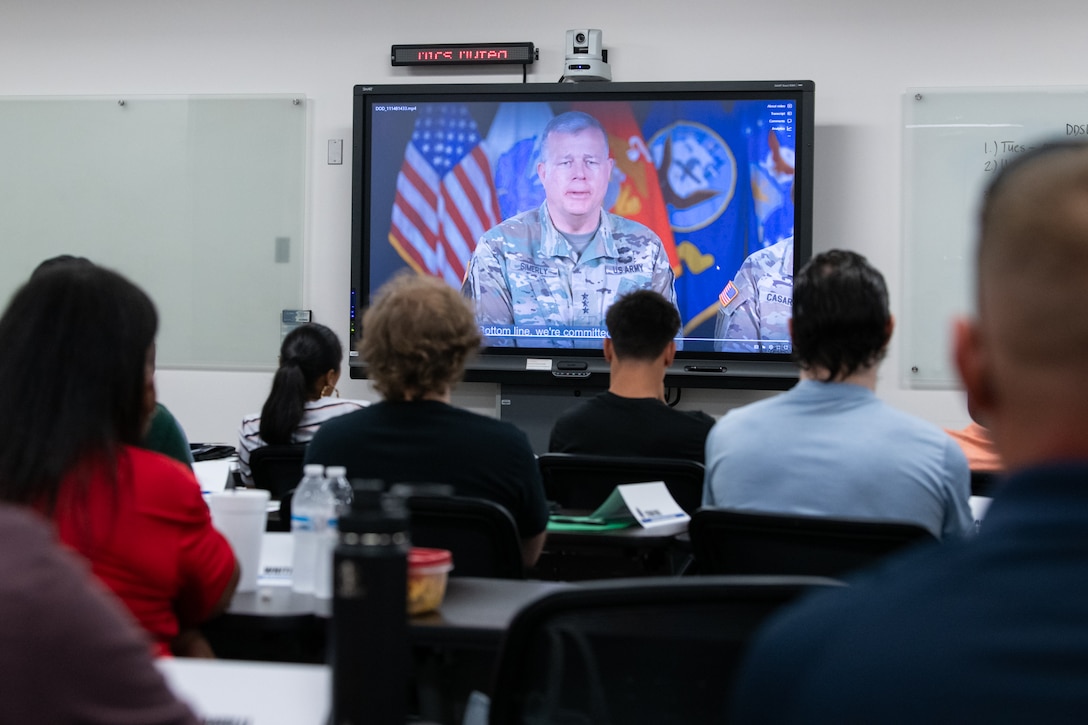 A group of people sit in a conference room looking at a tv screen with a video of a man wearing a camouflage military uniform on it.