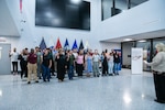 A large group of people stand in a building lobby in front of flags.