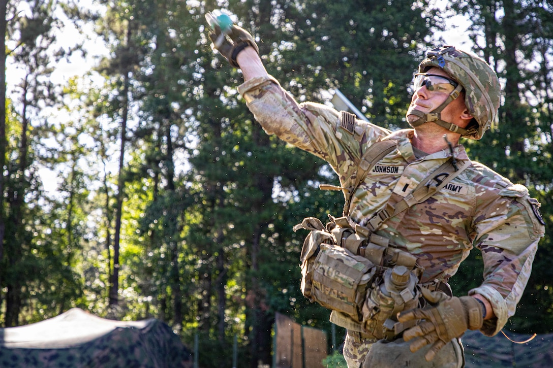 A soldier wearing a camouflage uniform and helmet throws a grenade in a forest setting during daytime, with two canopies in the background.