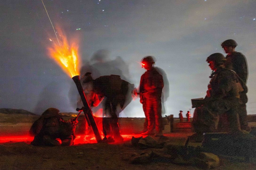 Marines partly illuminated by red light fire a mortar, which shoots flames into a dark, starry sky.