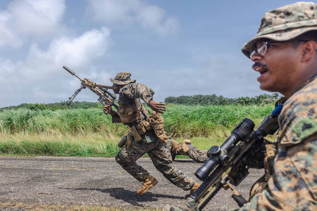 A Marine runs on a road next to a field while holding the pistol grip of a rifle with his right hand. Another Marine with his mouth open, in the foreground, looks left. Clouds and a blue sky are in the background.