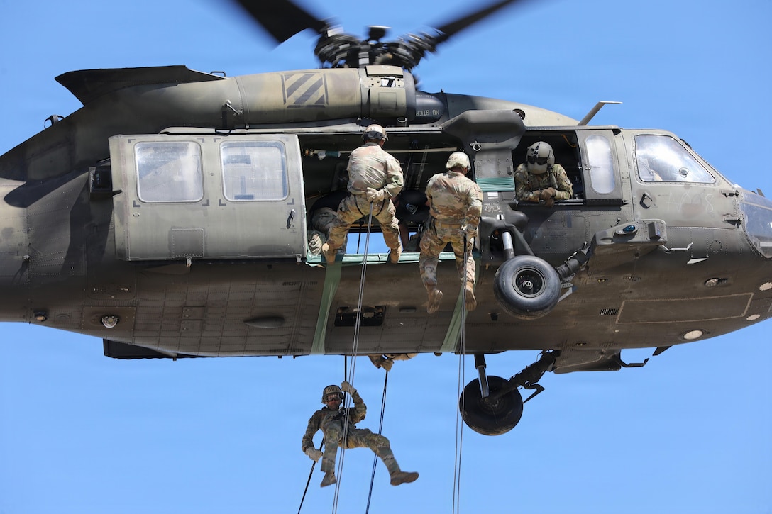 Three soldiers descend from a helicopter by rope while another inside the helicopter leans out a window to spot them.