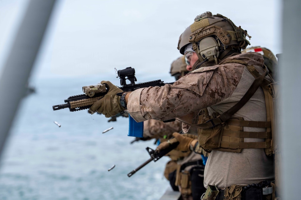 Two service members in camouflage uniforms fire rifles over the deck of a vessel. The shell casings fall toward the water below.