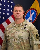 Army soldier dressed in combat uniform posing for a photo in front of the US flag and an organizational flag.