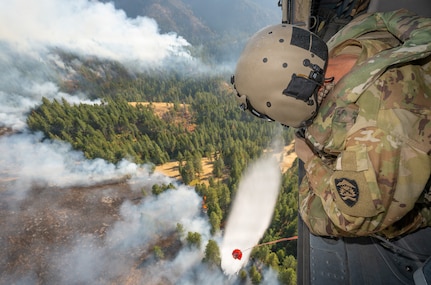 U.S. Army Staff Sgt. Kyle Sanders, a crew chief with G Company, 1st Battalion, 189th Aviation, watches as water releases from an HH-60M Black Hawk helicopter's bucket onto the Hagen Fire in Umatilla County, Oregon, Aug. 10, 2026.