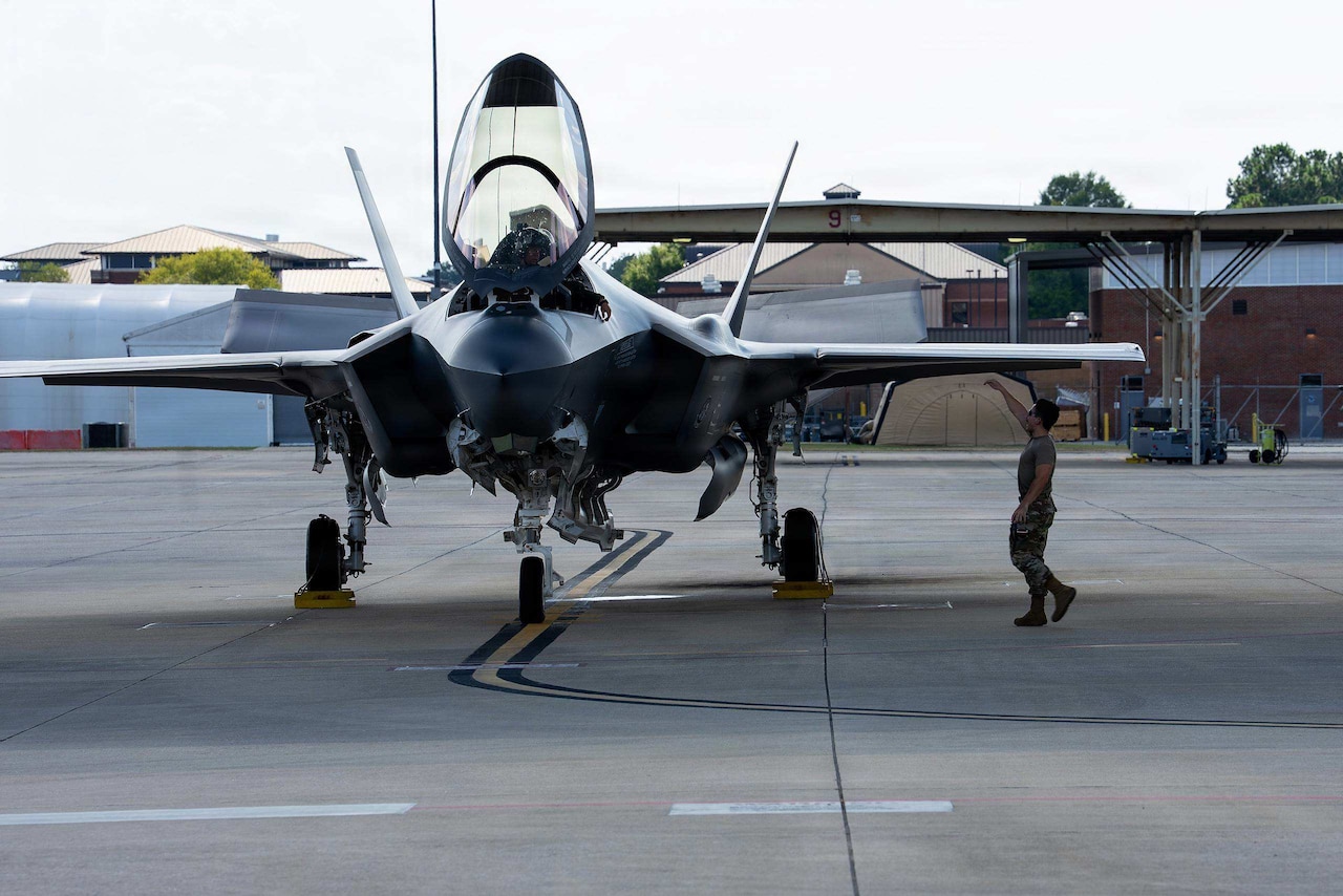 A man in a camouflage military uniform signals to a person in the cockpit of a military fighter jet parked on a tarmac.