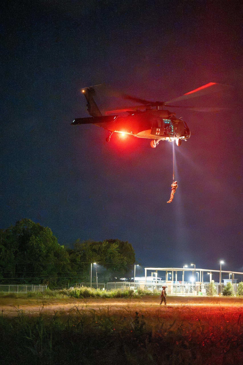 A military helicopter hovers in the air at night while hoisting a person in a camouflage military uniform into the aircraft.