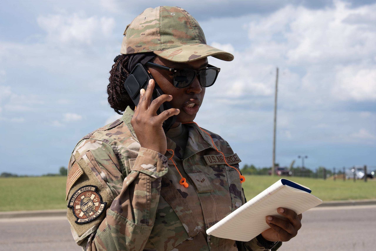 A woman in a camouflage military uniform speaks on a phone while looking at a notebook in her hand.