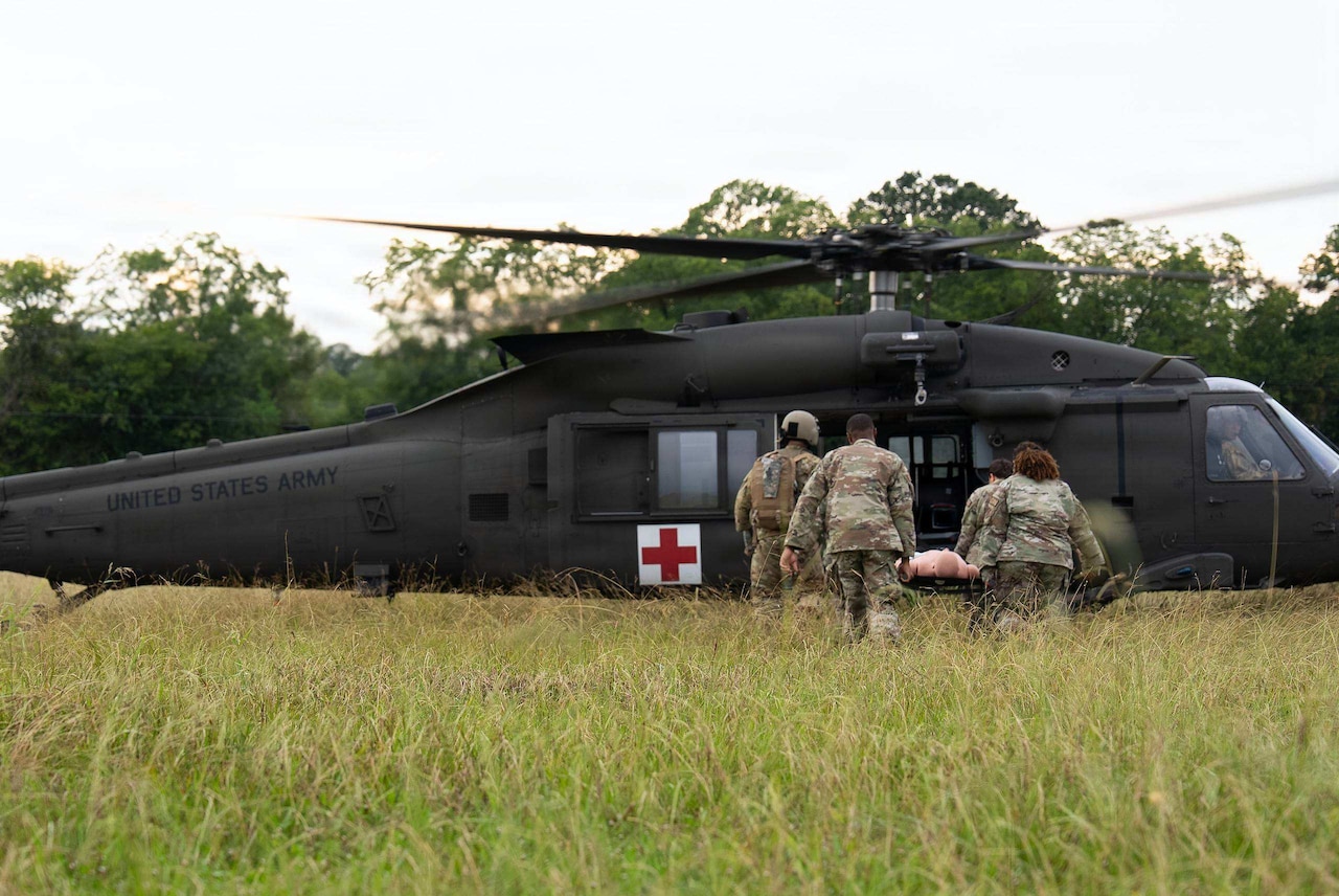 Four people in camouflage military uniforms carry a mannequin on a stretcher to a military helicopter parked in a grassy field.