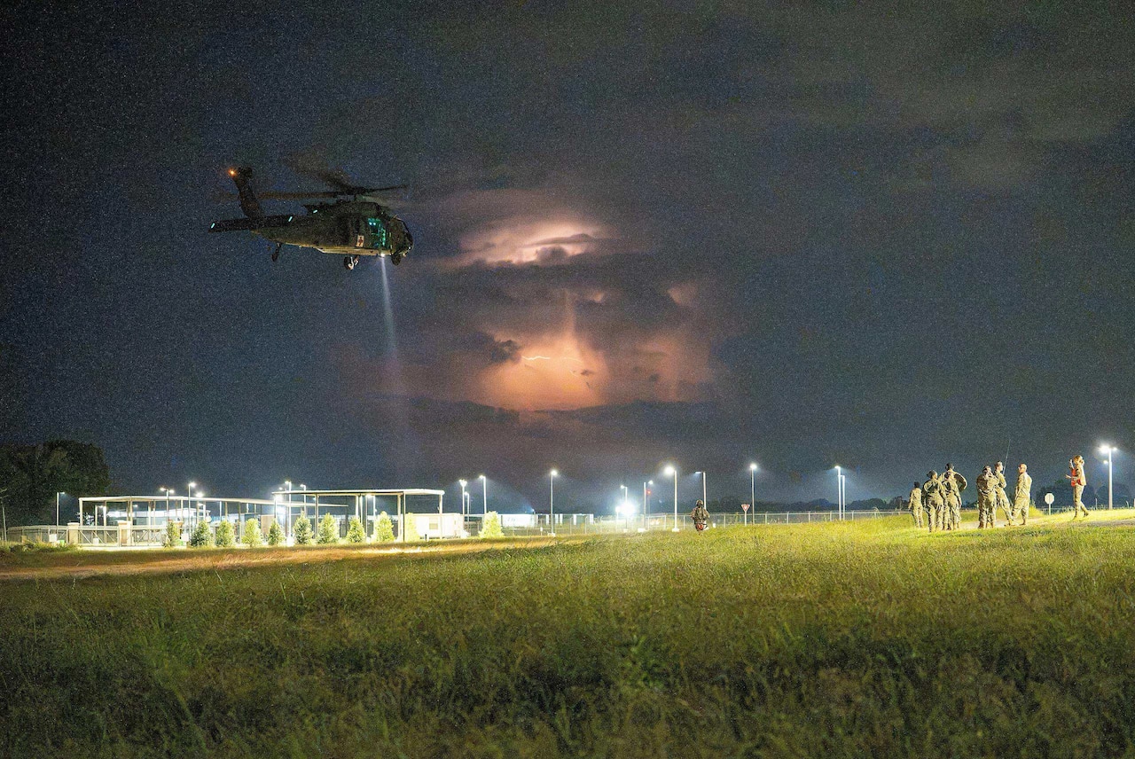 A military helicopter flies over an open field at night as eight people in camouflage military uniforms observe from the ground; a thunderstorm is in the background.