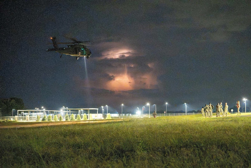 A military helicopter flies over an open field at night as eight people in camouflage military uniforms observe from the ground; a thunderstorm is in the background.