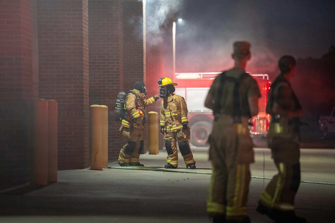 Two people in firefighter uniforms and gas masks speak outside a smoky building as two other people look on.