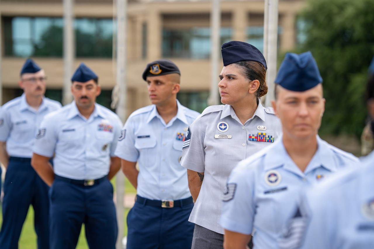 A guardian stands with other service members in formal uniforms in a line outdoors during daytime, with a building in the background.