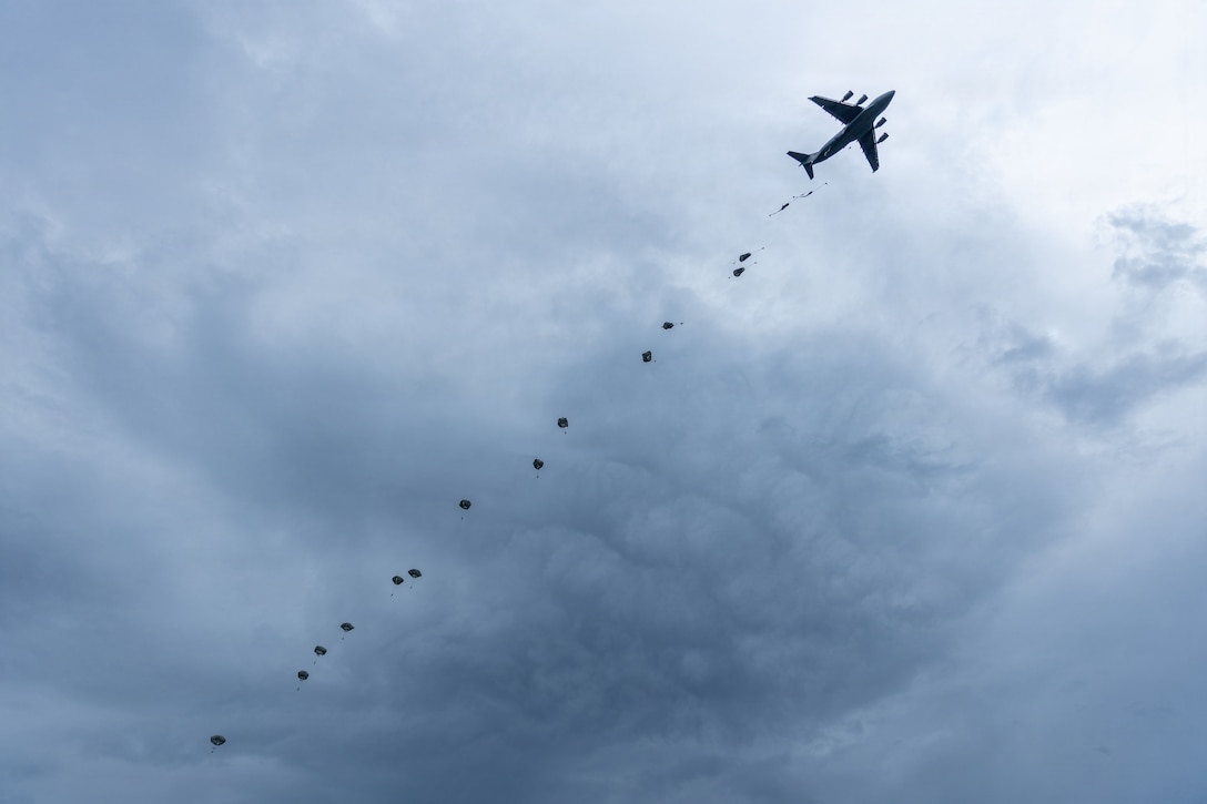A trail of 15 parachuters fall from the blue, cloudy sky behind an airplane.
