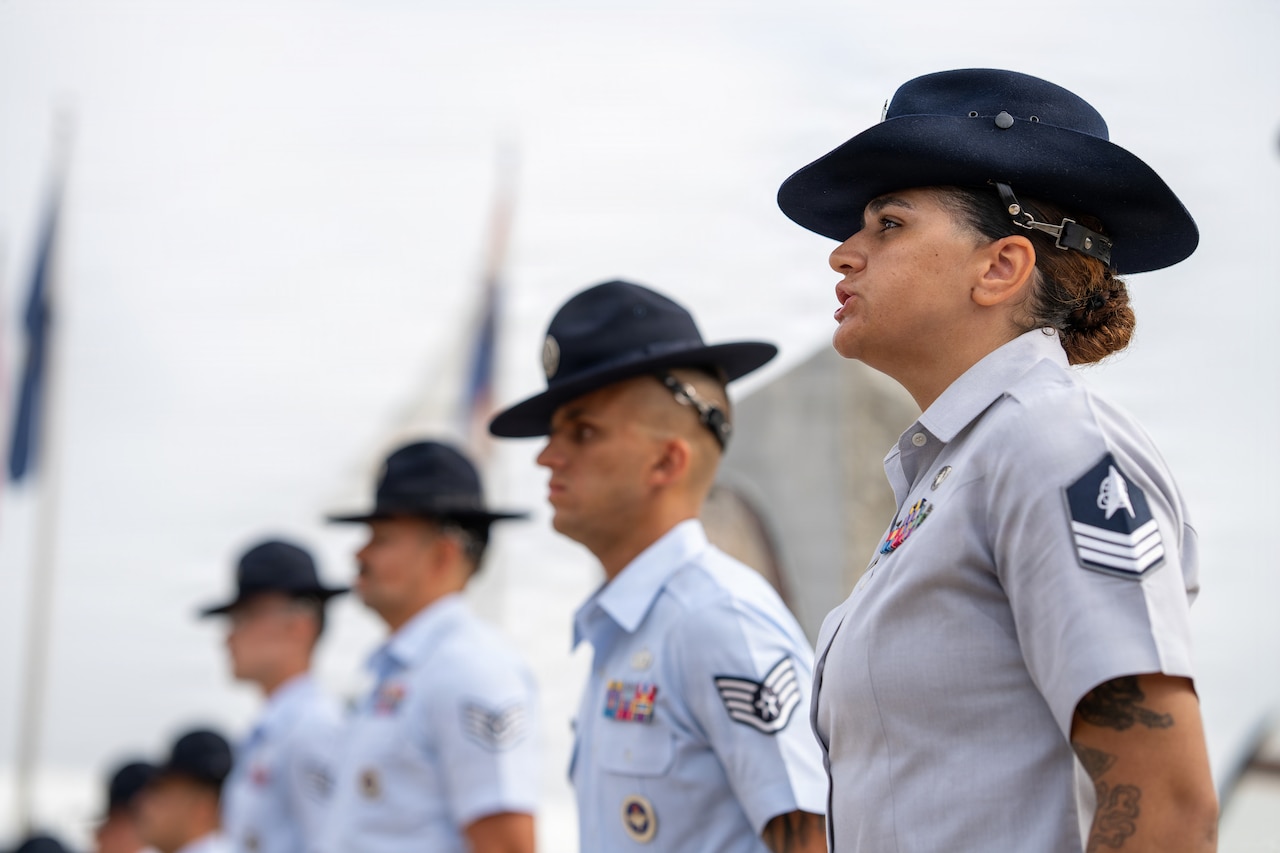 A guardian standing in a row with other service members speaks outdoors, with two flags on poles in the background.