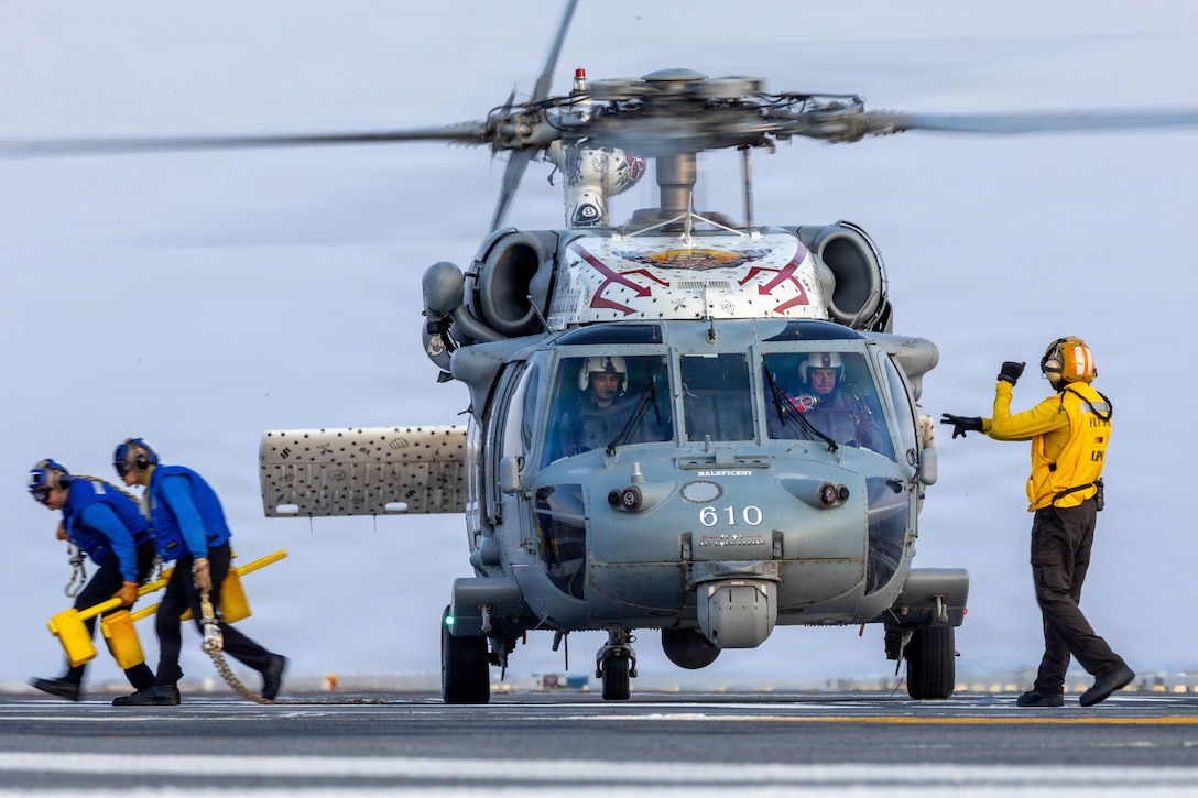A sailor gives signals to two sailors sitting in a helicopter on the deck of a ship, as two other sailors hunch down under the rotor while walking away.