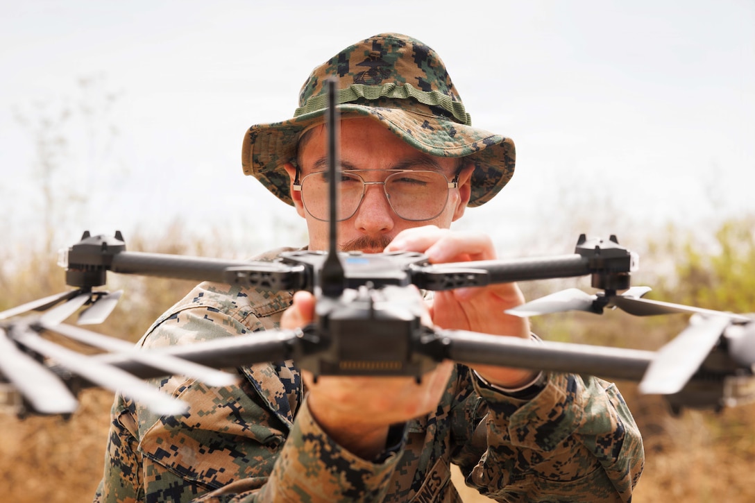 A Marine wearing a camouflage uniform and hat holds a small drone in front of him with both hands and closes one eye to focus on the craft outdoors during daytime.