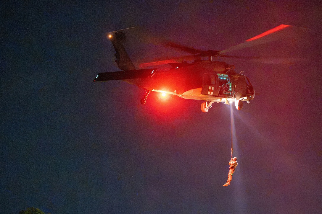 A service member attached to a cable ascends toward the open door of a hovering helicopter illuminated by red light in dark conditions.