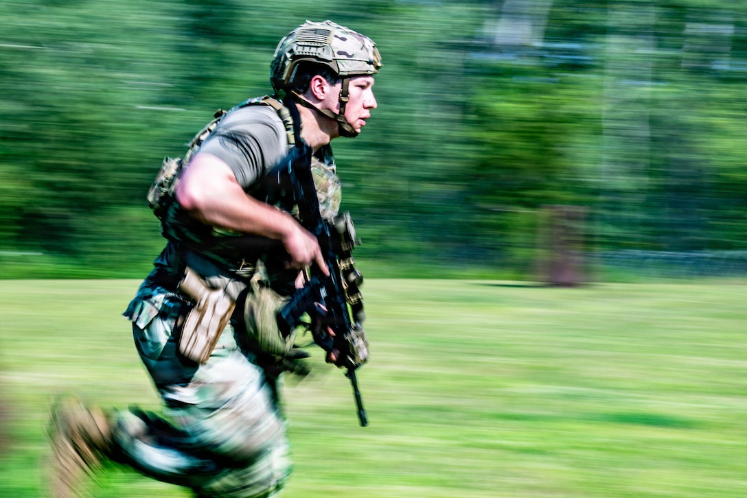 An airman wearing camouflage pants, a green T-shirt and tactical gear and carrying a rifle runs during daytime on a grassy field, with blurred trees in the background.