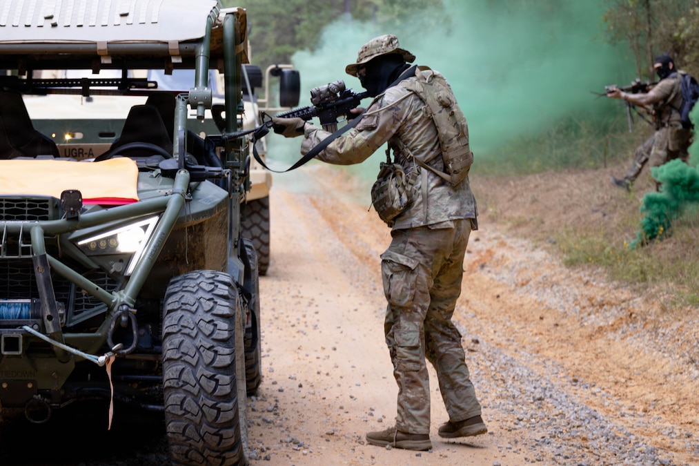 A soldier in a camouflage uniform and face mask points a rifle into a vehicle on a dirt road as another walks toward a vehicle behind the first, with green smoke wafting nearby.