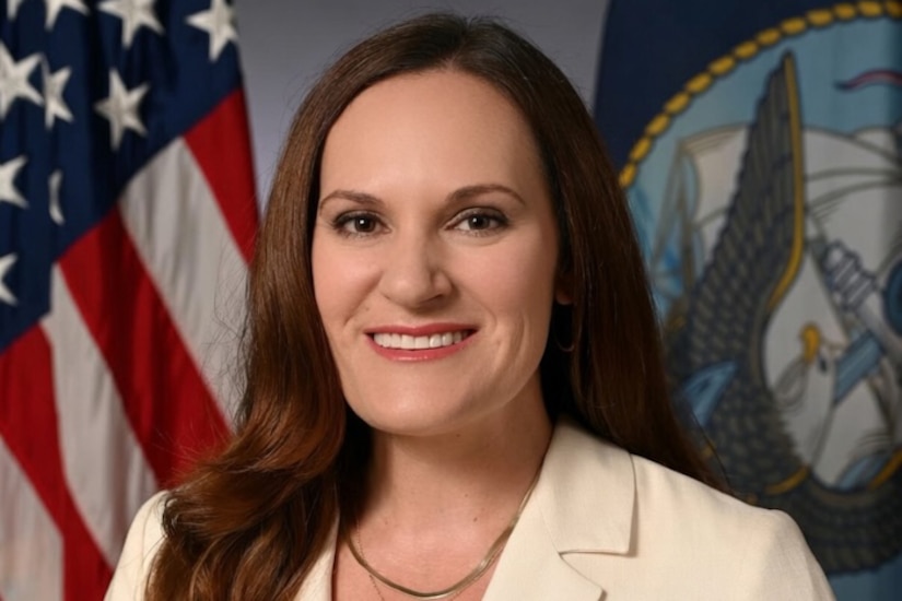 A woman in business attire poses for a portrait; behind her are the American flag and the U.S. Navy flag.